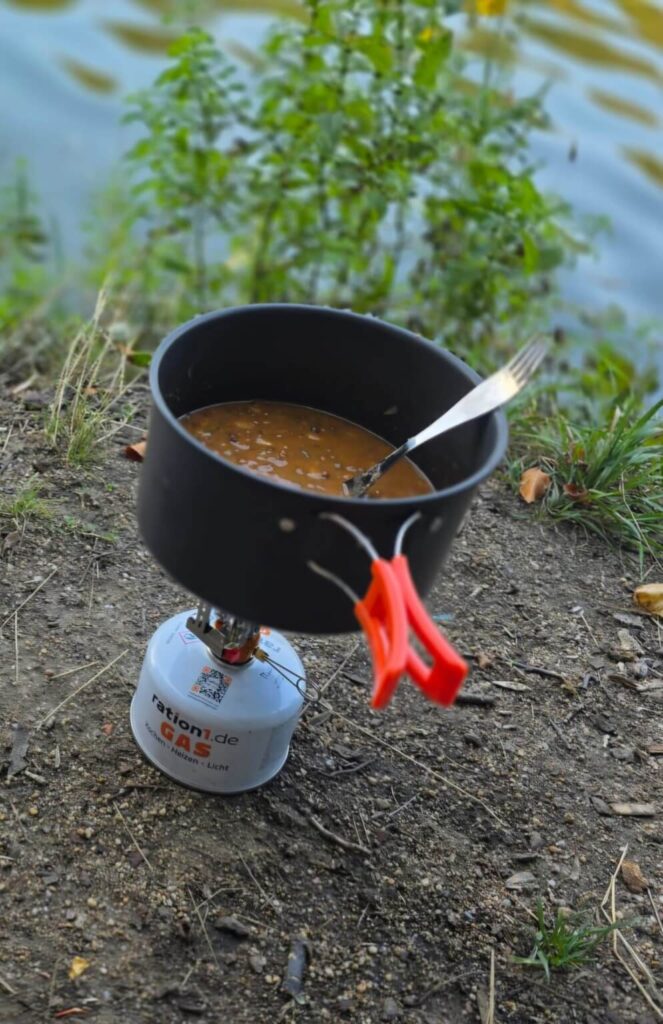 Gaskocher mit Topf und Essen am Ufer während einer Trekkingtour